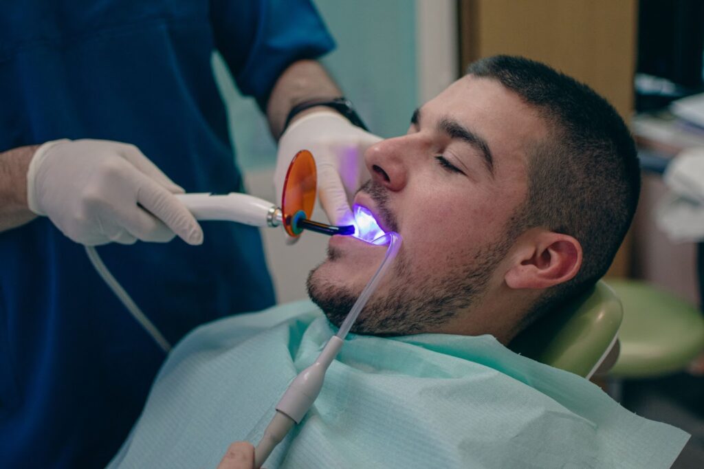 Man with eyes closed receiving dental treatment, wearing a teal bib, as a gloved professional uses a curing light tool inside his mouth. can I sue my dentist